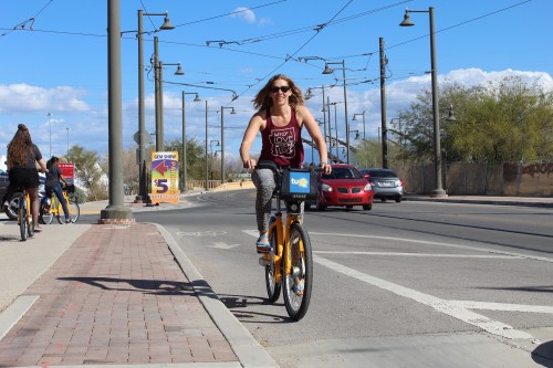 A woman rides a bike on a sunny street, smiling, with cars and other cyclists in the background.