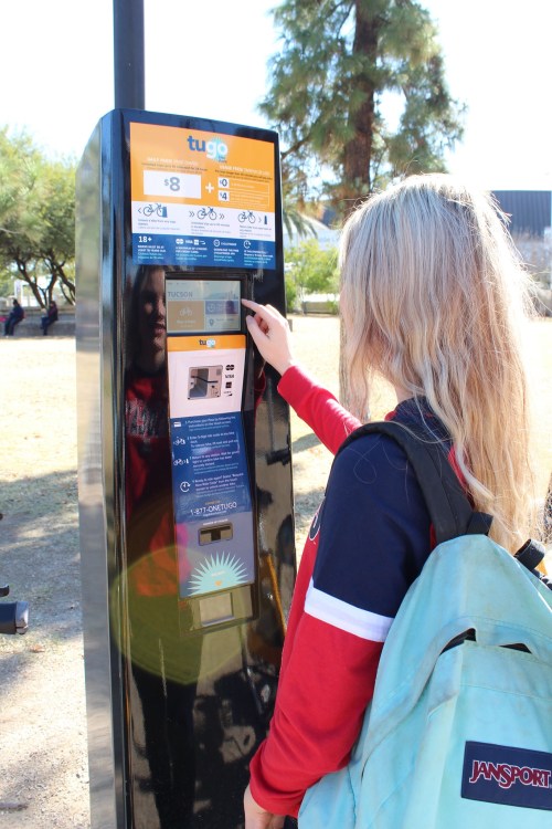 A person with long hair interacts with a bike rental kiosk in a park, wearing a backpack.