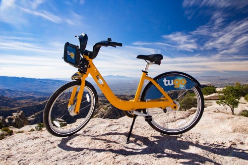 A yellow bike with a blue logo parked on a rocky surface, overlooking a scenic mountain view under a clear sky.