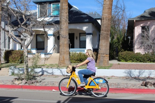 A woman rides a yellow bike past a house with palm trees and a clear blue sky.