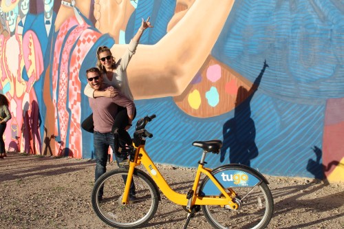 A man and woman pose playfully in front of a colorful mural, with a yellow bike nearby.
