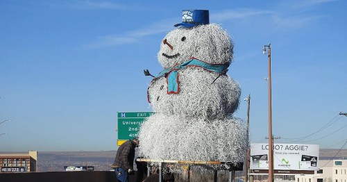 A large snowman made of wire and lights, wearing a blue hat and scarf, stands beside a highway.