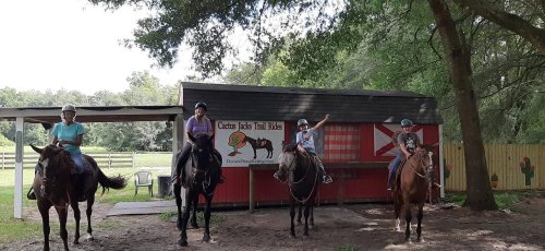 Four riders on horseback in front of a red barn, with a sign for Cactus Jacks Trail Rides in a wooded area.