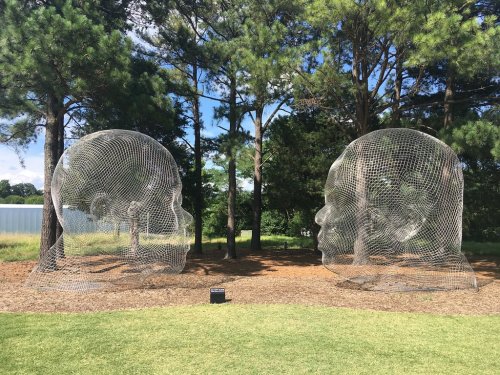 Two large wireframe sculptures of human heads facing each other, surrounded by trees and grass.