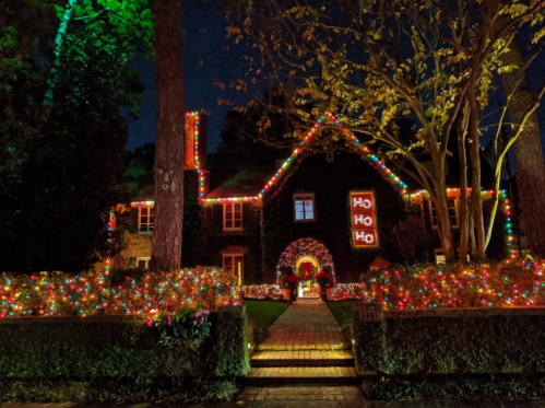 A festive house decorated with colorful Christmas lights and a "Ho Ho Ho" sign, surrounded by greenery at night.