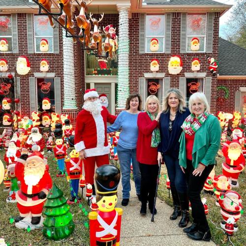 A festive holiday scene with four women posing with a Santa figure among numerous colorful decorations and lights.