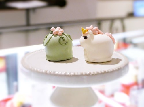 Two cute, decorative cakes on a stand: one green with flowers, the other a white unicorn with a golden horn.