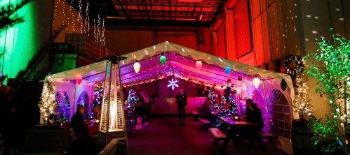 A festive outdoor tent decorated with lights, trees, and holiday ornaments, set in a warehouse environment.