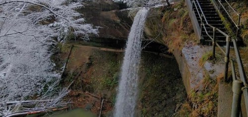 A waterfall cascading down a rocky cliff, surrounded by snow-covered trees and a staircase nearby.