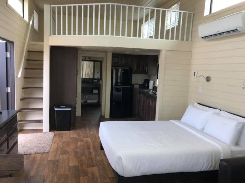 Cozy interior of a small cabin featuring a bed, kitchenette, and a loft area with stairs. Bright and inviting atmosphere.