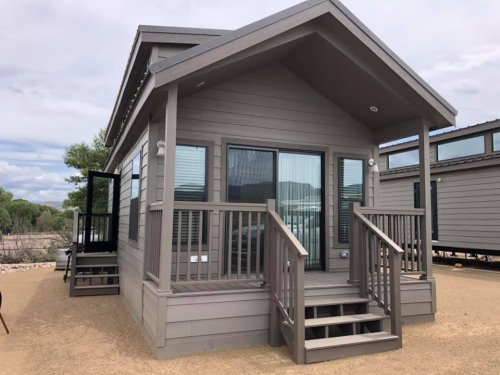 A modern, gray tiny house with a porch and steps, set in a natural outdoor environment.