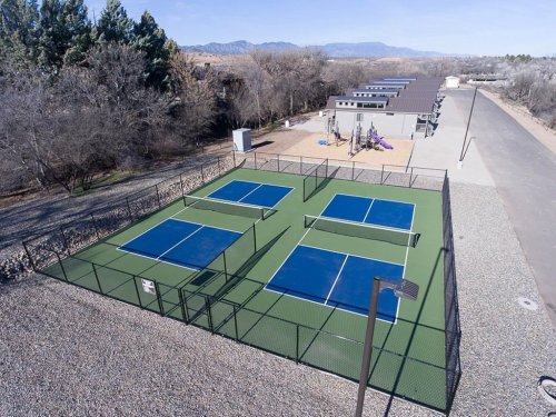 Aerial view of four pickleball courts surrounded by a fence, with a playground and buildings in the background.