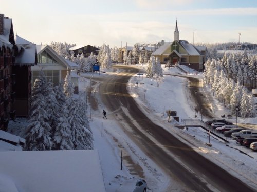 A snowy landscape with a winding road, a church, and buildings surrounded by frosted trees under a clear sky.