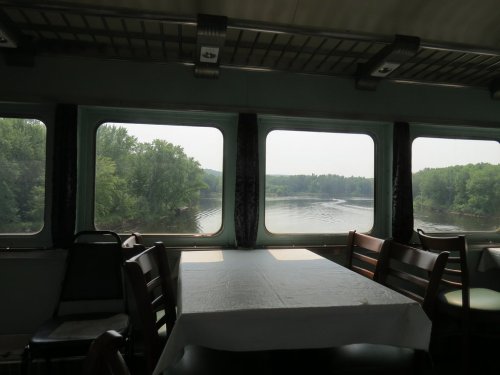 View from inside a boat, showing a table and windows overlooking a calm river surrounded by greenery.