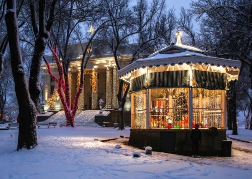 A snowy park scene featuring a decorated gazebo and a building adorned with lights, surrounded by bare trees.