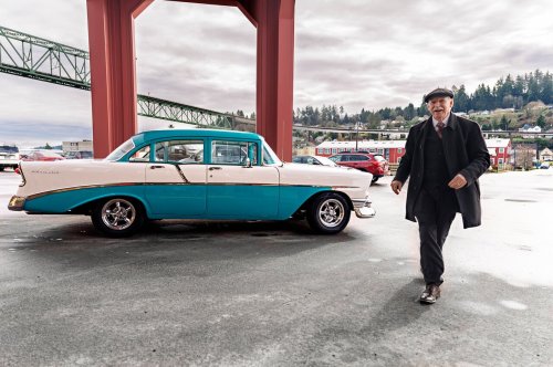 A man in a suit and cap walks past a vintage blue and white car under a bridge, with a scenic background.