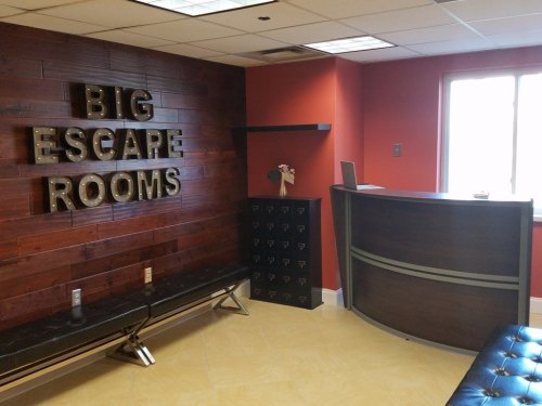 Interior of an escape room lobby featuring a wooden wall with "BIG ESCAPE ROOMS" sign, a reception desk, and seating.