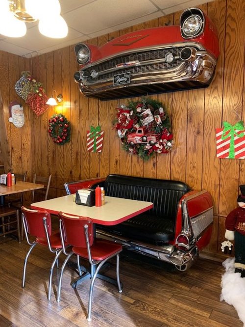 A retro diner interior featuring a vintage car front on the wall, festive decorations, and a booth with red chairs.