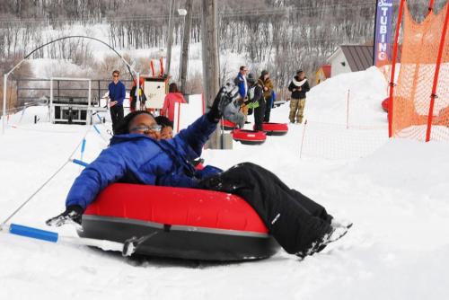 A person in a blue jacket enjoys tubing down a snowy slope, waving with excitement. Snowy landscape and other tubers in the background.