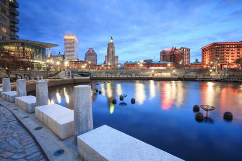 City skyline at dusk reflecting in a calm river, with illuminated buildings and a stone walkway in the foreground.