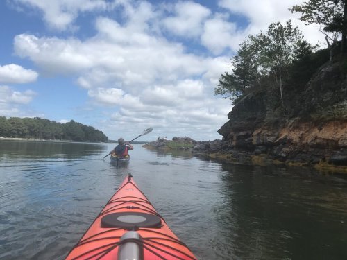 A person kayaking on calm water, with rocky shores and trees under a partly cloudy sky.