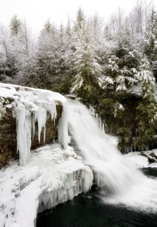 A snowy waterfall cascades over icy rocks, surrounded by frosted trees in a winter landscape.