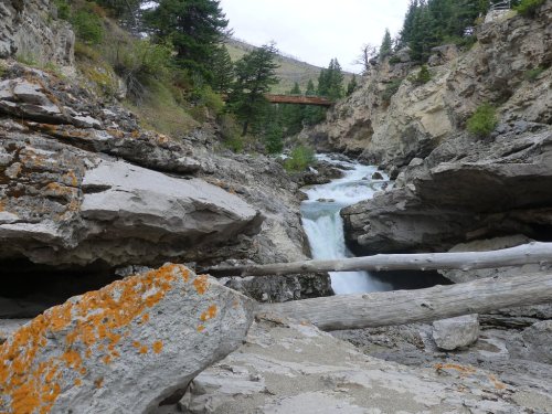 A rocky riverbed with flowing water, surrounded by trees and a wooden bridge in the background.