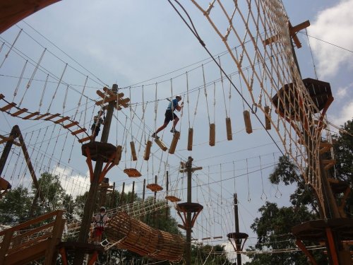 A person navigates a high ropes course, balancing on wooden planks suspended between wooden structures against a blue sky.