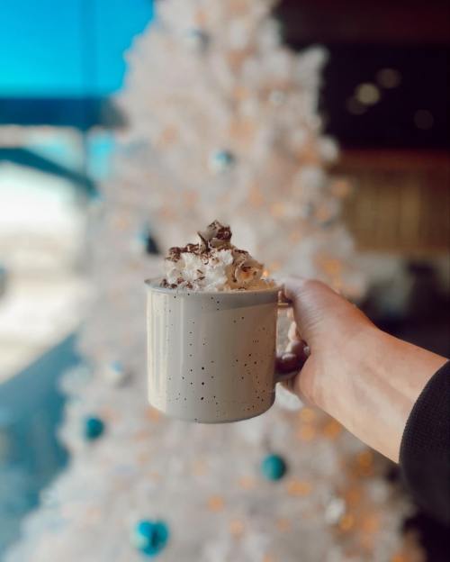 A hand holding a mug topped with whipped cream and popcorn, with a blurred white Christmas tree in the background.