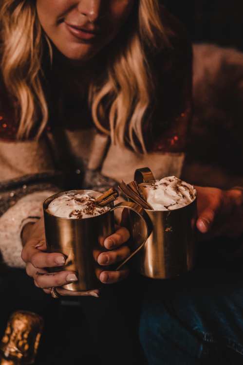 A person holds two brass mugs filled with whipped cream and garnished with cinnamon sticks, smiling softly.