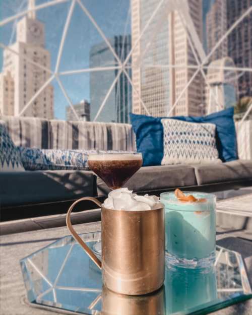 Three colorful cocktails on a table with a city skyline in the background, featuring a cozy lounge setting.