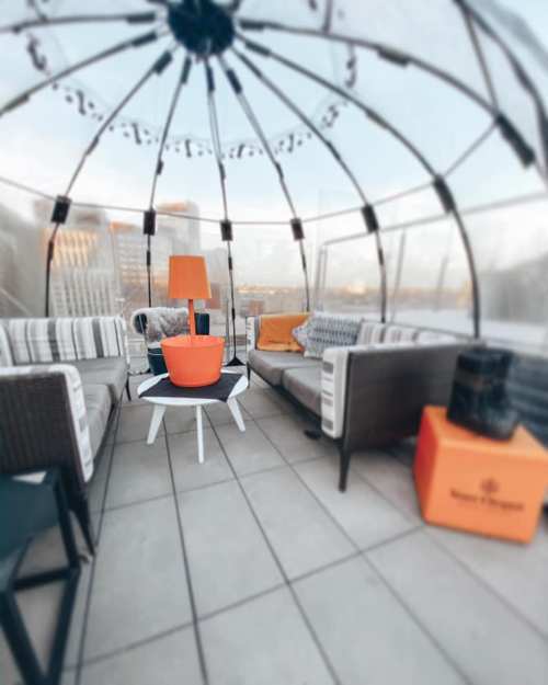 A cozy rooftop lounge with a transparent dome, featuring stylish seating and an orange lamp on a table.