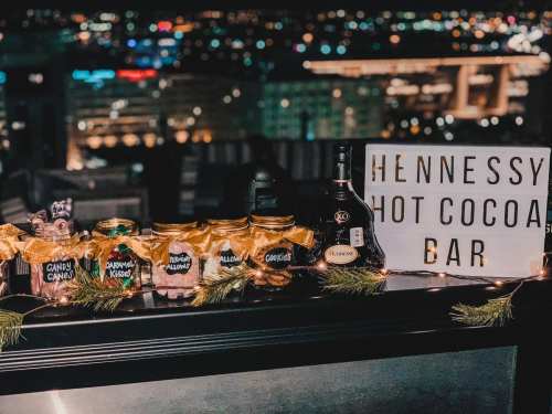A festive hot cocoa bar with jars of toppings, a Hennessy bottle, and a sign, set against a city skyline at night.