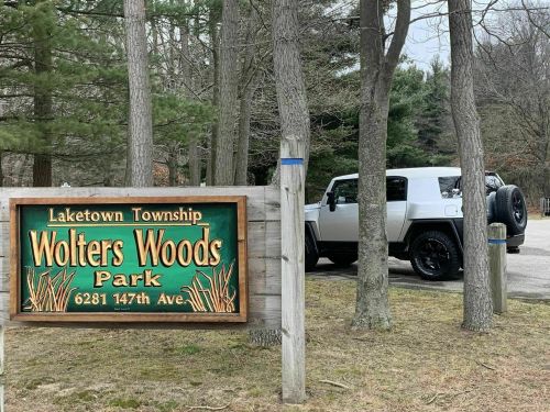 A white vehicle parked near a sign for Wolters Woods Park in Laketown Township, surrounded by trees.