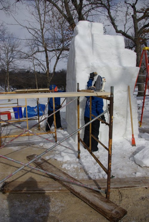 Two people working on a large snow sculpture, using scaffolding and tools in a snowy outdoor setting.