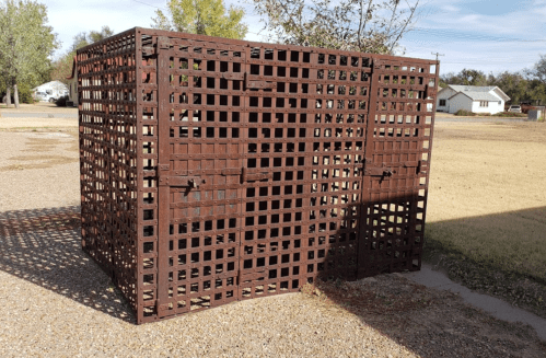 A large, brown metal cage with a grid pattern, positioned outdoors on a gravel surface.