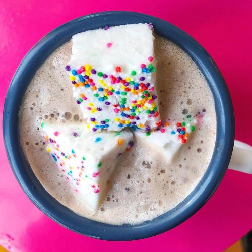 A mug of hot chocolate topped with colorful, sprinkle-covered marshmallows on a bright pink surface.