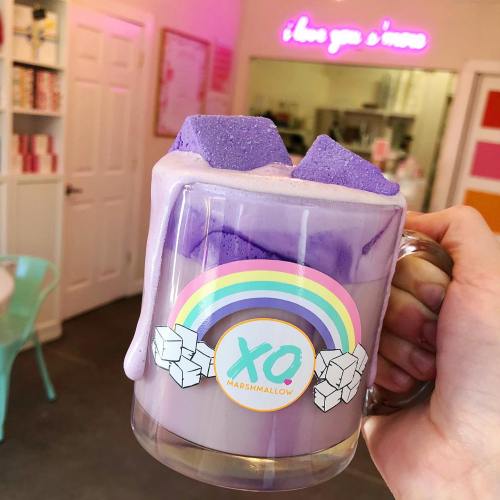 A hand holds a clear mug filled with purple drink topped with marshmallows, set in a colorful café.
