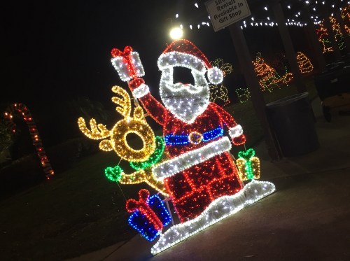 A brightly lit Santa Claus figure holding a gift, accompanied by a reindeer, surrounded by festive holiday lights.