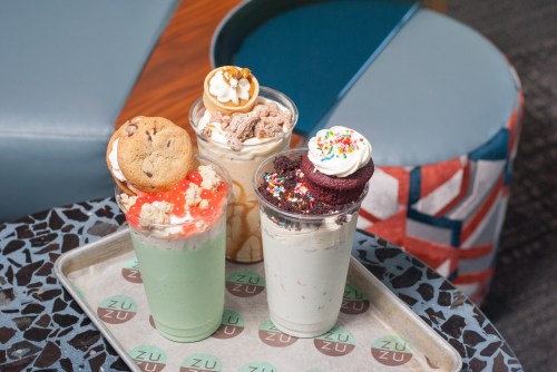 Three colorful milkshakes topped with cookies, whipped cream, and sprinkles, served on a tray.