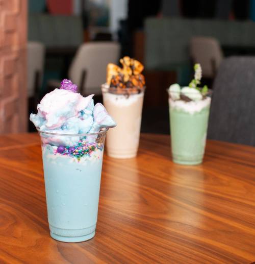 Three colorful milkshakes in clear cups on a wooden table, featuring whipped cream and toppings.