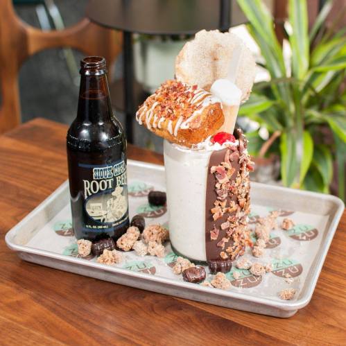 A decadent milkshake topped with a donut, whipped cream, and nuts, served with a bottle of root beer on a tray.