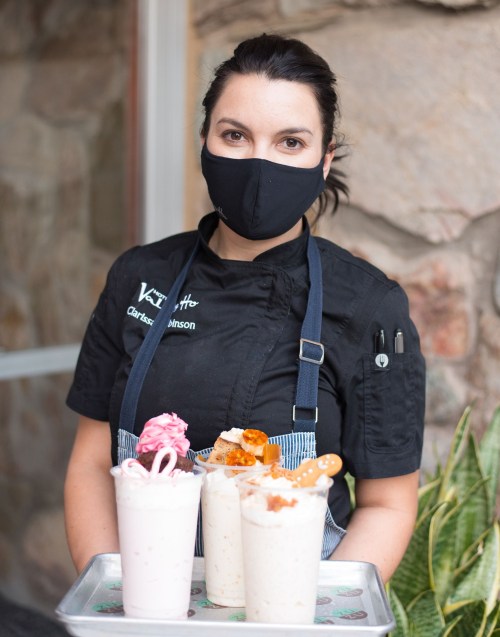 A chef in a black uniform and mask holds a tray with three colorful milkshakes, standing in front of a stone wall.