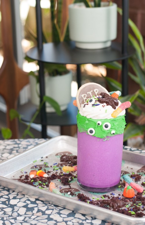 A colorful purple milkshake topped with green frosting, candy, and googly eyes, served on a tray with scattered treats.