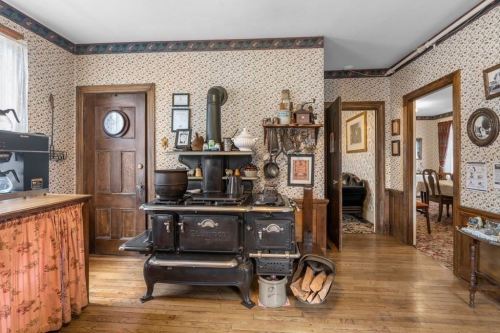A vintage kitchen with a black stove, floral wallpaper, wooden accents, and a cozy dining area visible in the background.