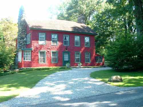 A red two-story house with stone accents, surrounded by greenery and a gravel driveway.