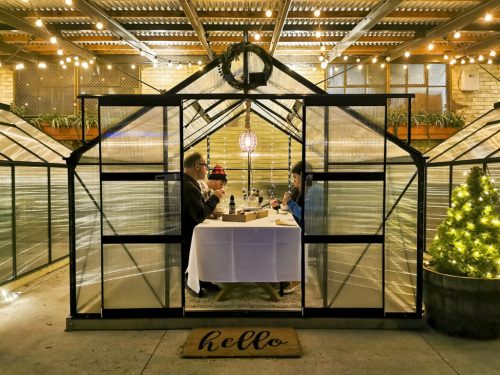 Two people dining inside a greenhouse-style structure, decorated with lights and a small tree, creating a cozy atmosphere.