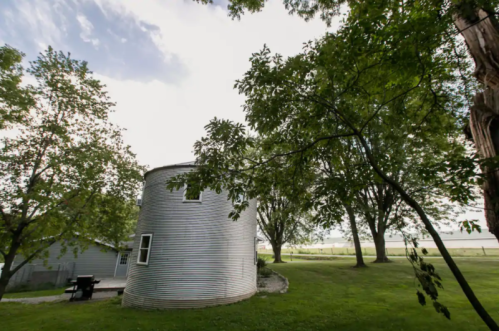 A round, metal silo surrounded by trees and green grass under a cloudy sky.
