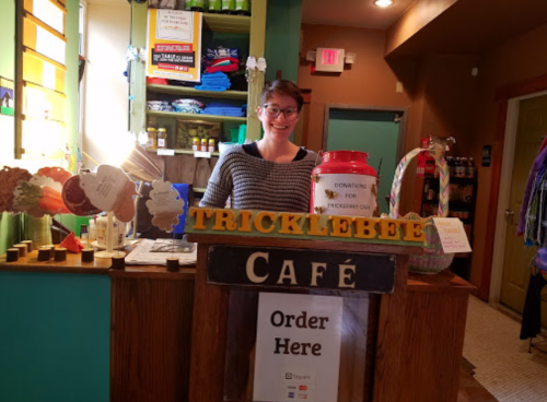A smiling barista stands behind the counter of Tricklebee Café, with a donation jar and colorful decor in the background.