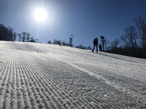 A sunny ski slope with groomed snow, skiers in the distance, and ski lifts visible against a clear blue sky.